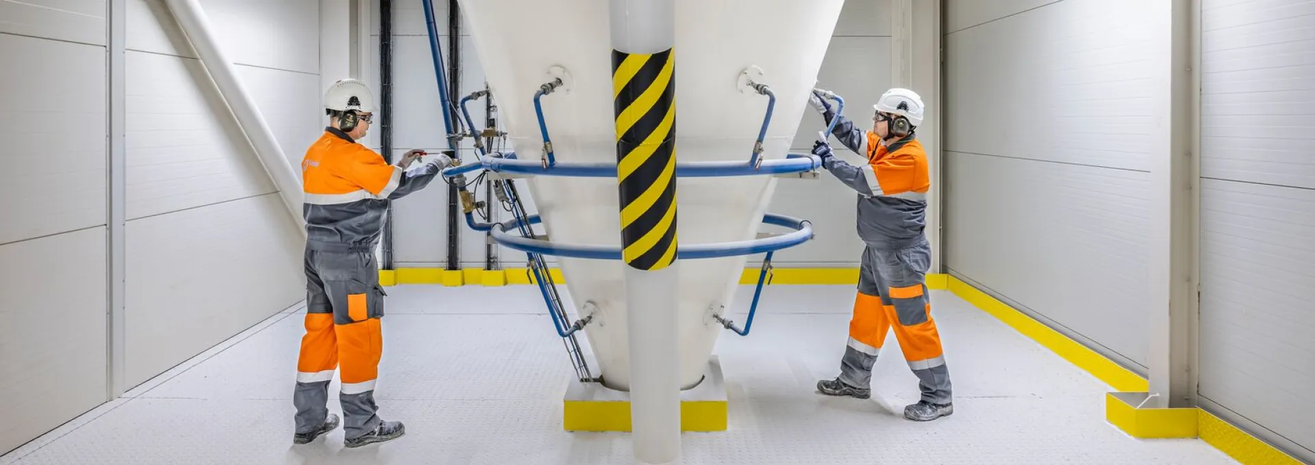 Two operators in protective equipment monitoring and operating a large industrial storage silo inside a processing facility.