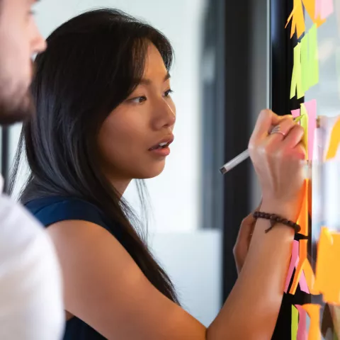 A focused Asian businesswoman mentor head coach write an idea or task on post sticky notes on verr wall