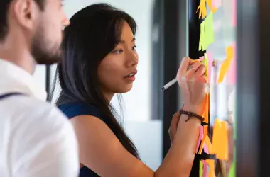 A focused Asian businesswoman mentor head coach write an idea or task on post sticky notes on verr wall