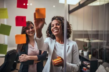 Creative businesswoman writing on sticky notes on a glass wall, colleague looking