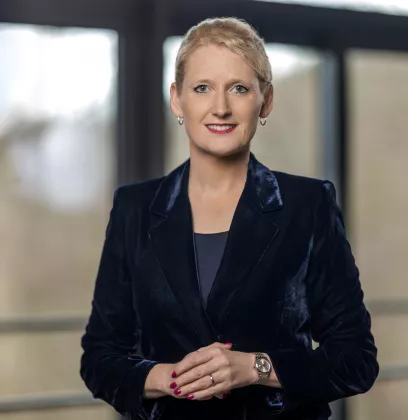 Business professional wearing a dark velvet blazer and watch, standing in a modern office environment with large windows and natural light.