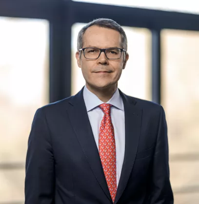 Corporate executive wearing a dark suit and red patterned tie, standing in a bright office environment with large windows and natural light.