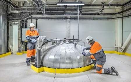 Two workers wearing PPE inspect and maintain a large industrial vessel inside a production facility with visible piping, cabling and process equipment.